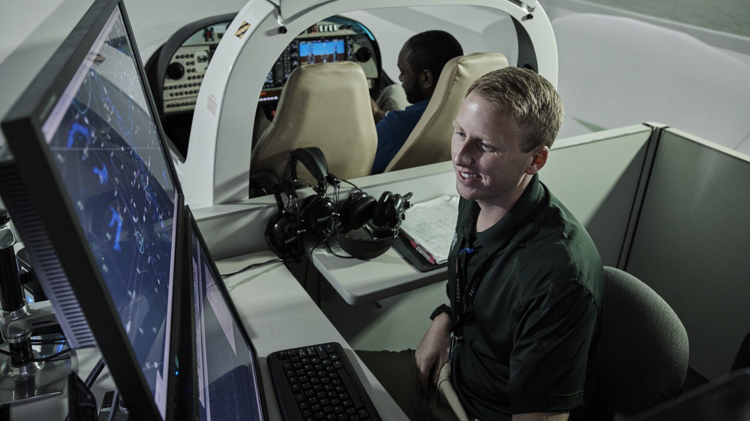 Student looking at computer screens, monitoring air traffic, while another student is in the flight simulator behind him.